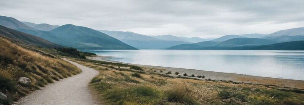 A quiet lake below low mountains under an overcast sky, with a gravel path running along the shore