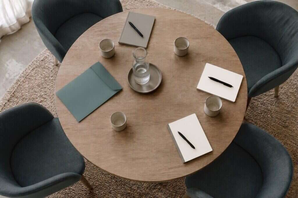 A round table seen from above, set for four with notebooks, pens, cups and a carafe of water, ready for a session to begin